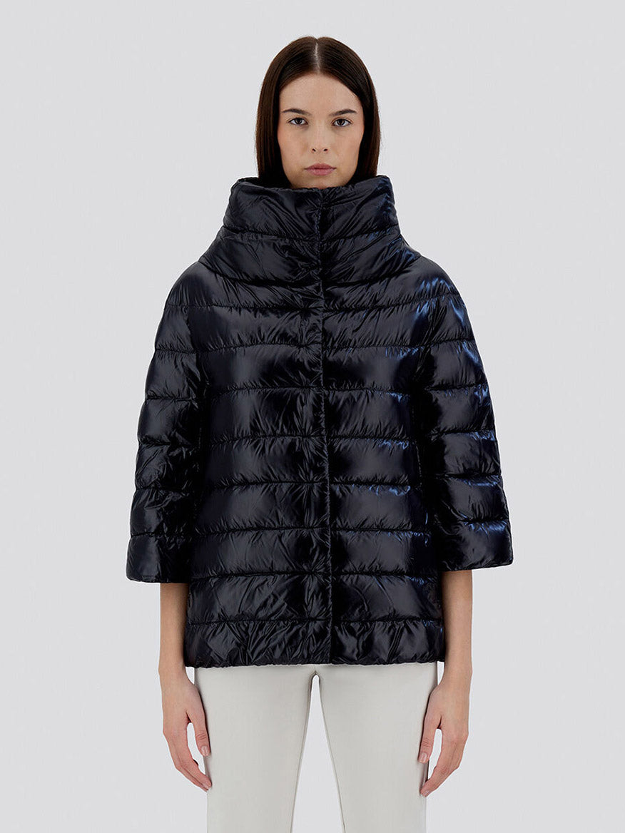 A woman stands facing forward, wearing the Herno Aminta 3/4 Sleeve Cocoon Jacket in Black. Its shiny, ultralight nylon material complements the oversize fit and high collar, with three-quarter-length sleeves adding to its chic design. She pairs it effortlessly with white pants against a plain light gray background.