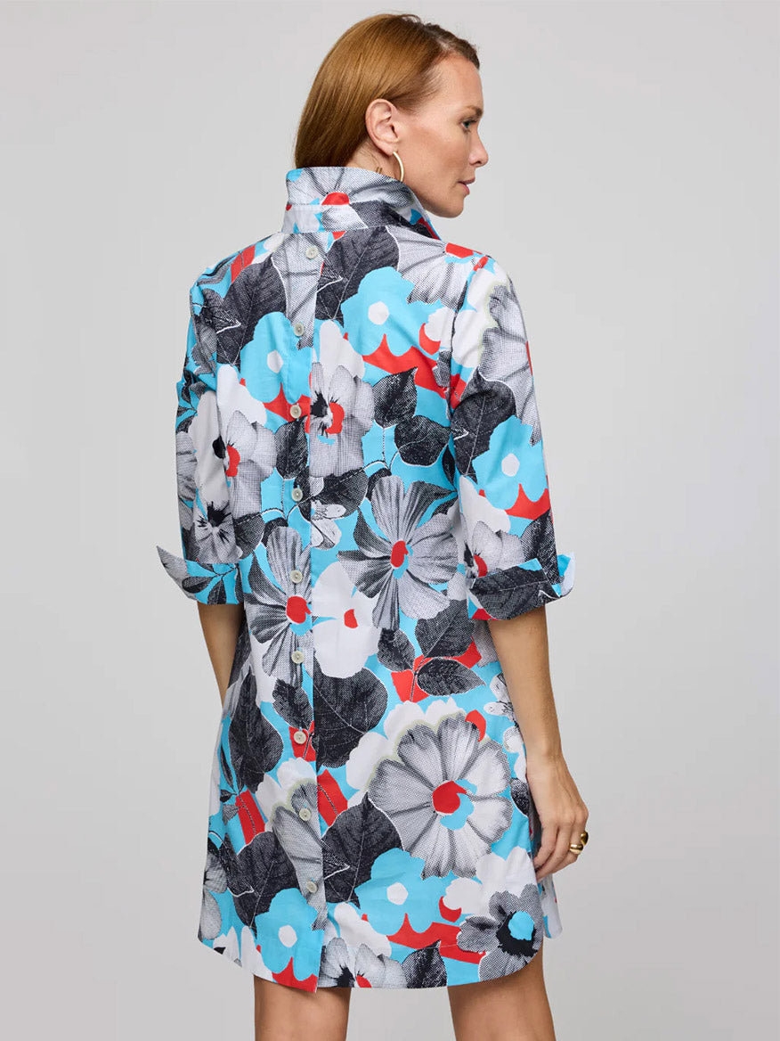 A woman stands facing away, wearing the Hinson Wu Aileen 3/4 Sleeve Pop Art Flower Dress in Blue Combo, featuring knee-length refined cotton and exclusive large blue, red, white, and black floral prints with sleeves rolled to the elbows.