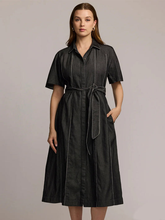 A woman models the Hinson Wu Joanna Short Sleeve Denim Dress in Black Wash, featuring a belted waist, button-down front, and white vertical stitching, made from a tencel cotton blend against a plain background.