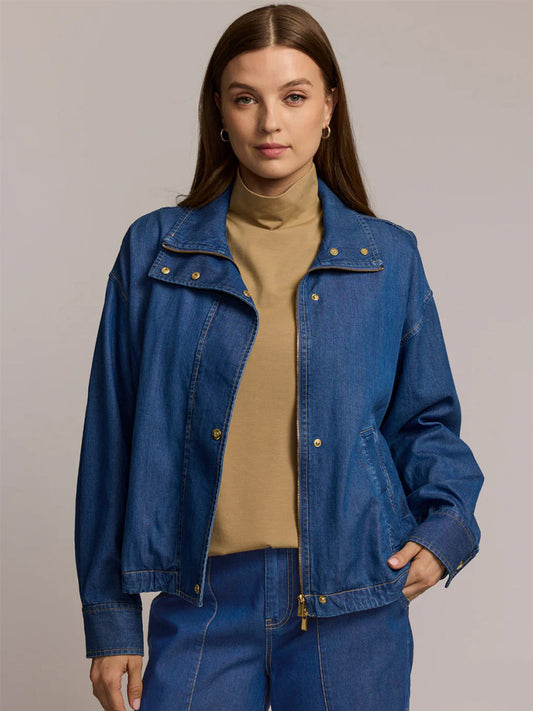 A woman wears the Hinson Wu Louise Long Sleeve Denim Jacket in Medium Wash over a tan turtleneck and matching blue denim pants, standing facing the camera with one hand in her pocket.