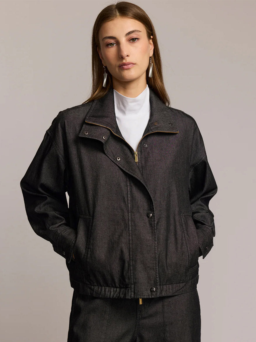 A woman with straight brown hair wears the Hinson Wu Louise Long Sleeve Denim Jacket in Black Wash over a white turtleneck, hands in pockets, standing against a plain background to showcase its effortless, refined style.