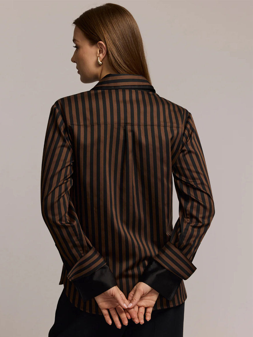 A woman with straight brown hair, seen from behind with hands clasped, wears the Hinson Wu Margot Long Sleeve Lace Print shirt in Caramel/Black—one of this season’s chicest looks.