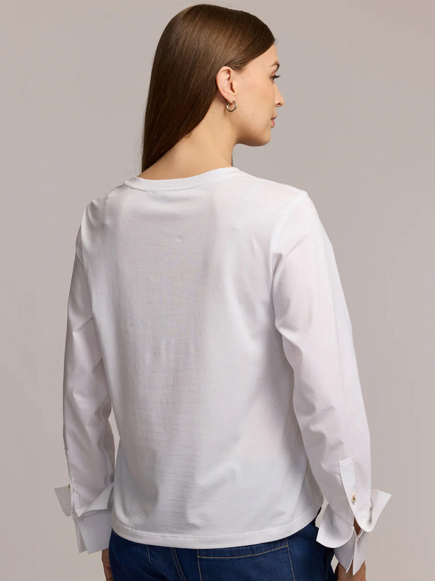 A woman with long brown hair stands with her back to the camera, wearing the Hinson Wu Reagen Long Sleeve Foundation Layer in White and blue jeans against a neutral background—an effortlessly chic look.