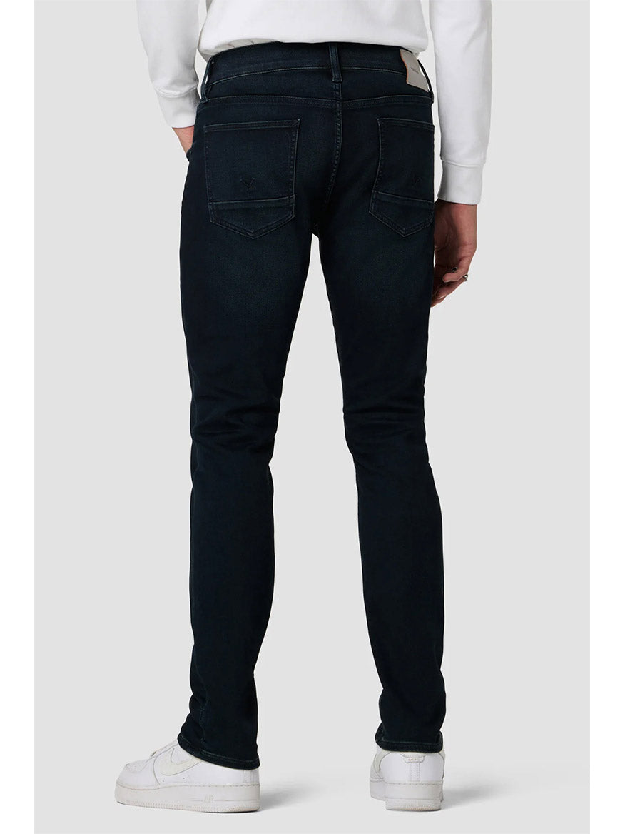 A person from the waist down viewed from behind wearing dark indigo Hudson Blake Slim Straight Jeans in Hayworth and white sneakers.