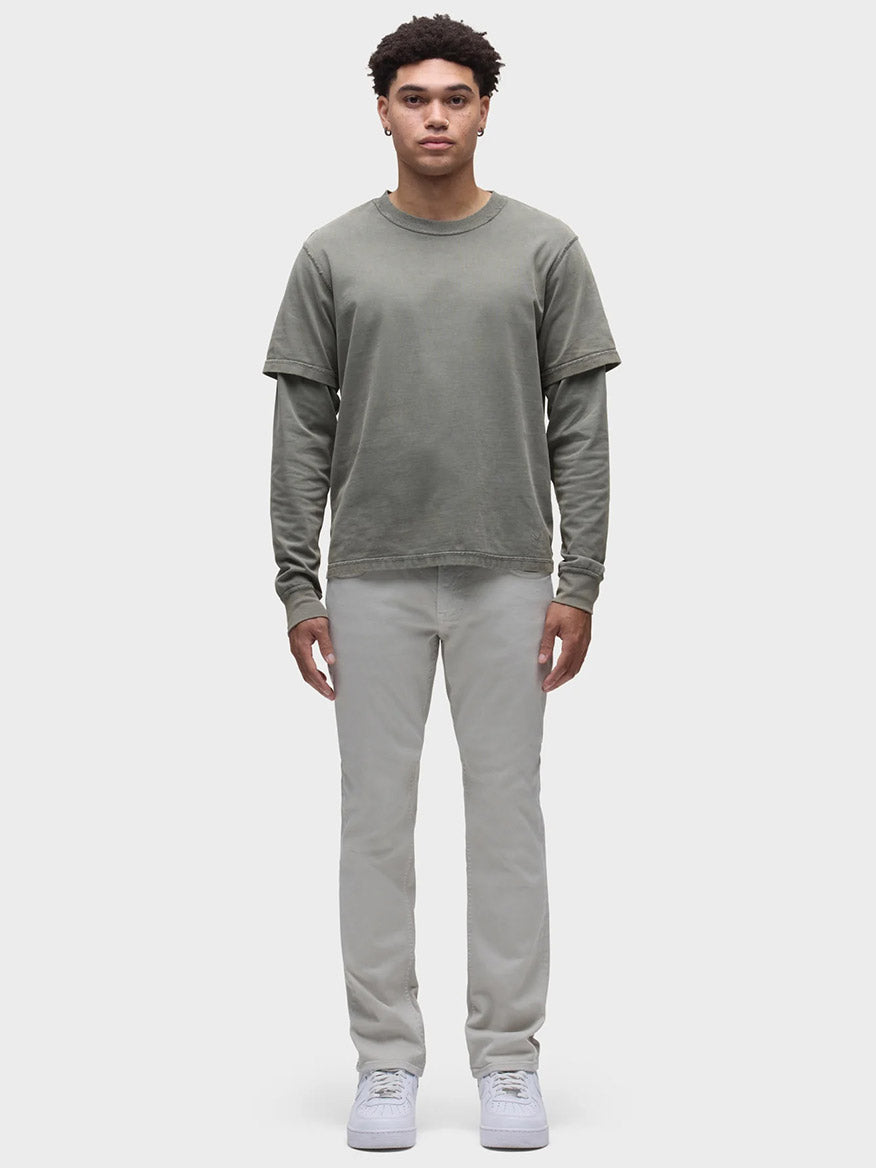 A person stands facing forward, dressed in a layered olive-green long-sleeve and short-sleeve shirt, paired with Hudson Blake Slim Straight Twill Pant in Ash and white shoes, against a plain white background.