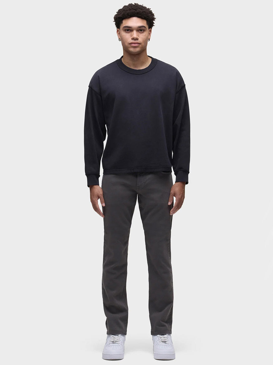 A person stands facing forward, wearing a dark sweater, Hudson Blake Slim Straight Twill Pant in Grey, and white shoes against a neutral background.