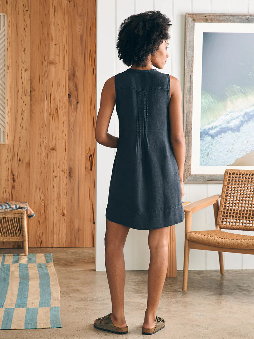 A woman in a sleeveless Faherty Brand Isha Basketweave Dress in Washed Black stands looking at a picture on the wall, in a room with wooden panels and a striped rug.