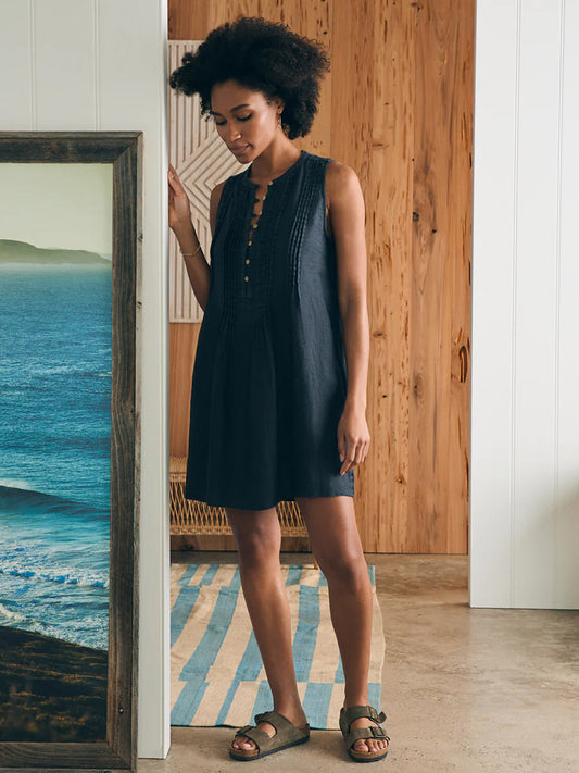 A woman in a sleeveless Faherty Brand Isha Basketweave Dress in Washed Black and sandals standing next to a mirror in a room with a wooden wall and a coastal painting.
