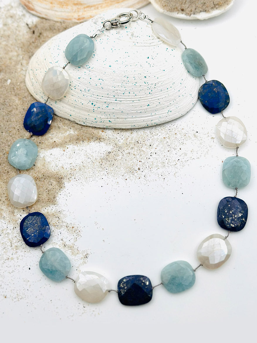 An 18" Flat Lapis, Aquamarine & Moonstone Necklace from JMNYC Designs is displayed on sand next to a large seashell.