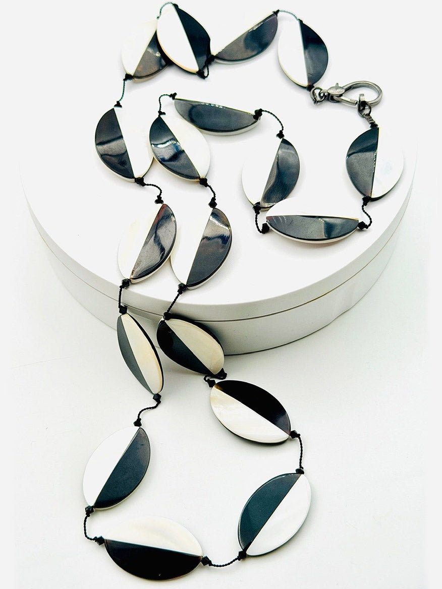 The JMNYC Designs 36" Oval Mother of Pearl and Black Tab Necklace showcases elegant oval beads split into bold black and white halves, all expertly strung on black thread and finished with a sleek metal clasp, beautifully presented on a white circular platform.