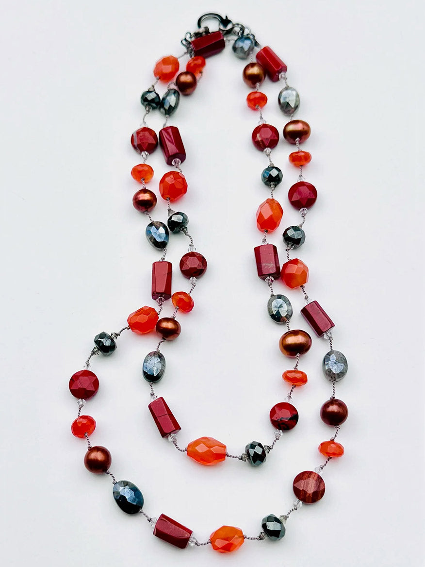 The JMNYC Designs 36" Red Jasper, Carnelian & Silverite Mix Necklace showcases an elegant double strand of alternating red, orange, brown, and silver beads in various shapes and sizes, beautifully arranged against a white background. This striking necklace is finished with a secure sterling silver clasp for added sophistication.