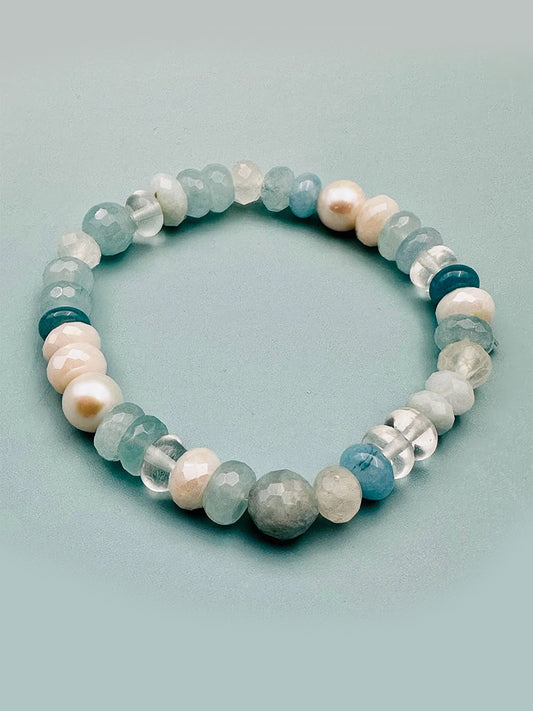The JMNYC Designs Aquamarine, Moonstone, Freshwater Pearl, & Crystal Quartz Stretch Bracelet features alternating beads and is shown on a light blue surface.
