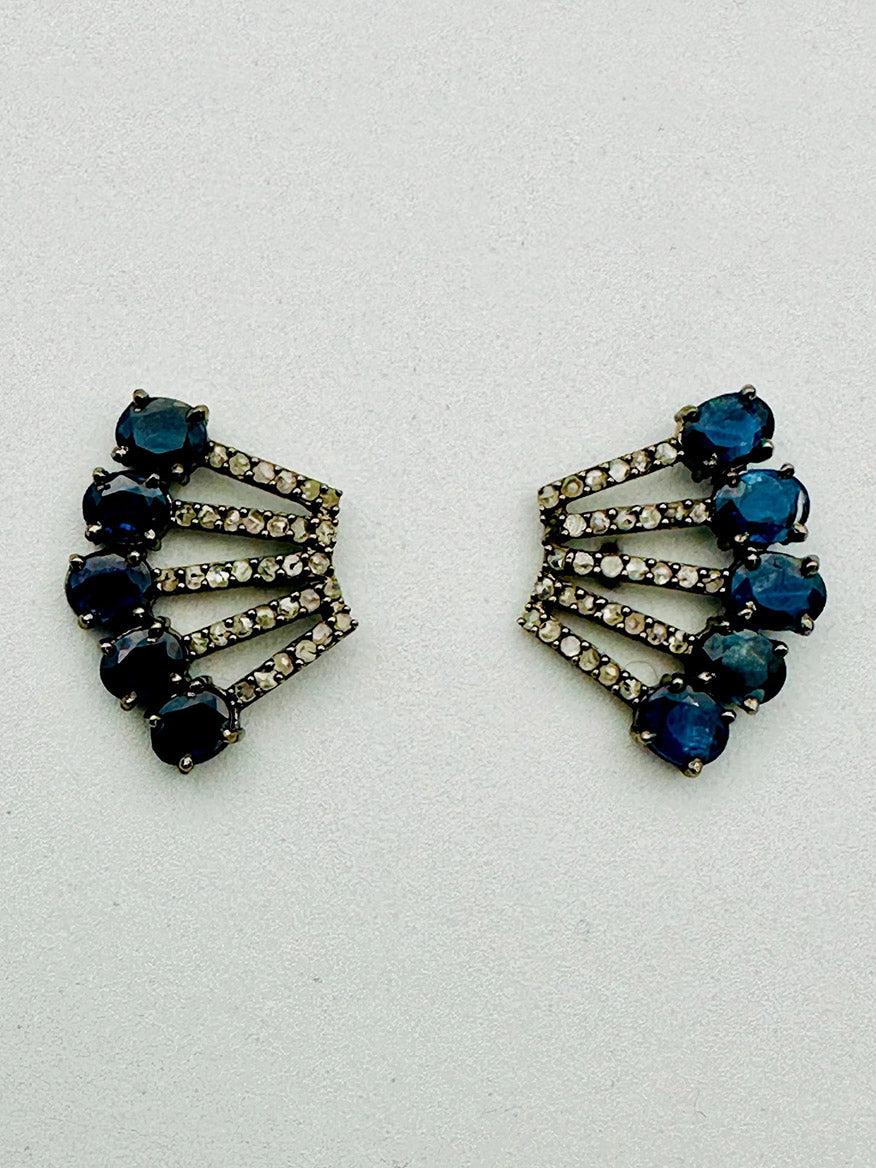 The JMNYC Designs Blue Sapphire Diamond Sterling Silver Post Earring features three rows of round blue sapphires and small clear stones in a fan-like, geometric design on a light sterling silver background.