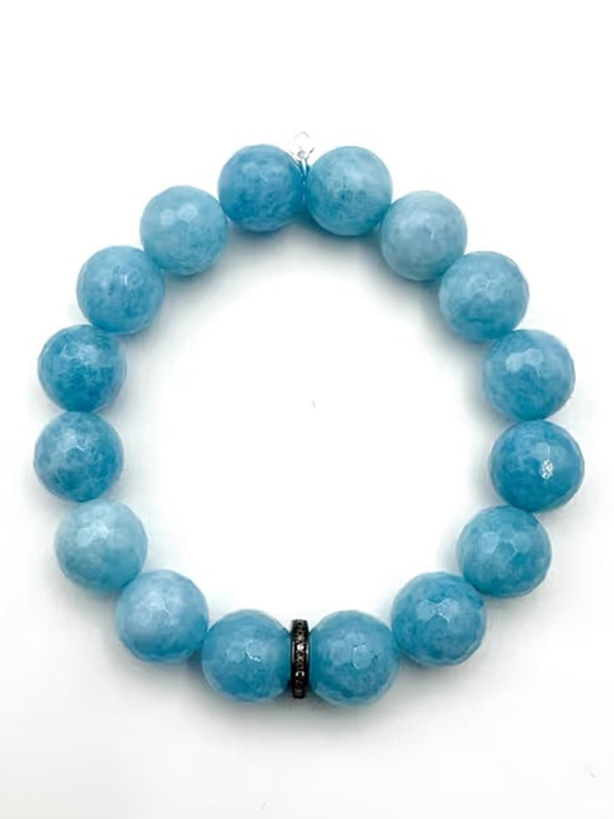The JMNYC Designs Light Blue Faceted Quartz Ball with Sterling Silver Diamond Rondelle is a stretch bracelet featuring round, faceted light blue quartz beads, highlighted by a single black accent bead and a sterling silver diamond rondelle, arranged in a circle on a white background.