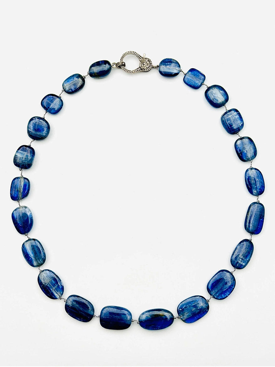 The JMNYC Designs Smooth Oval Kyanite Nugget Necklace, showcasing lustrous blue oval stones and finished with a Sterling Silver Diamond Clasp, is elegantly displayed in a circle on a plain white background.