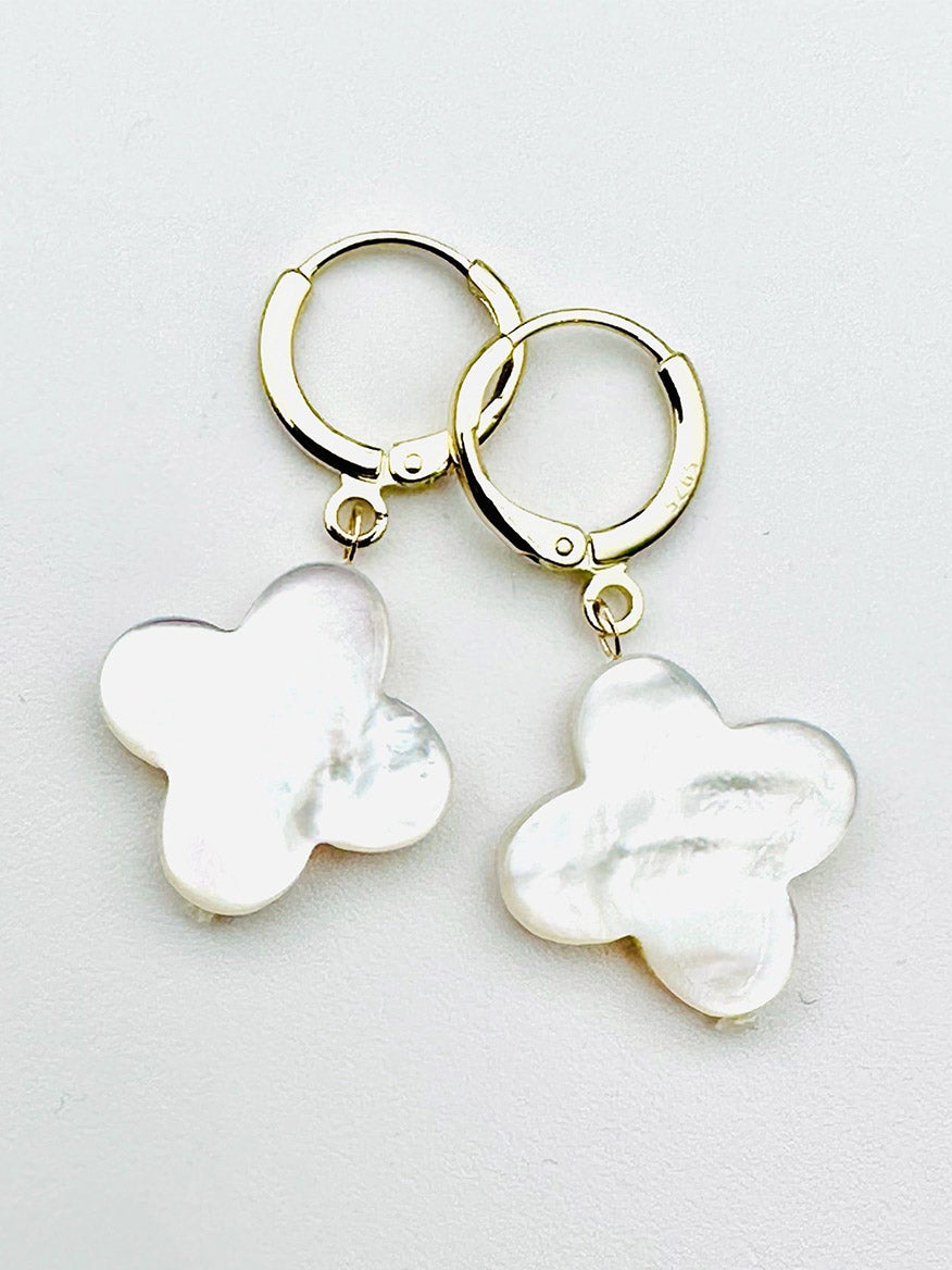 JMNYC Designs White Mother of Pearl Clover Gold Fill Huggie Earrings feature gold fill hoops with dangling, irregularly shaped white mother of pearl charms in a four-petal clover design, shown against a plain white background.