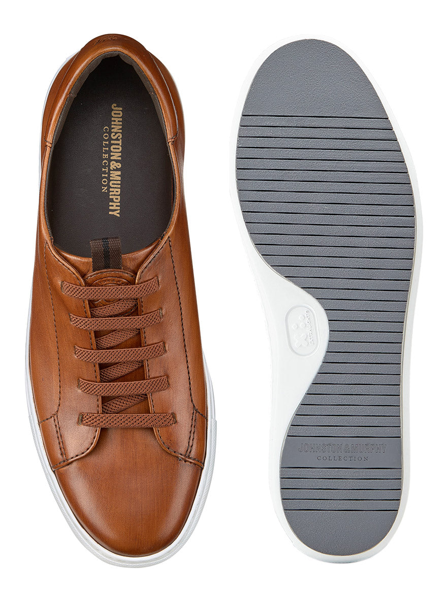 A pair of J & M Collection Anson Lace-to-Toe sneakers in hand-stained brown calfskin with white rubber soles is shown from above and below, featuring visible branding inside the shoe and highlighting comfort and craftsmanship.