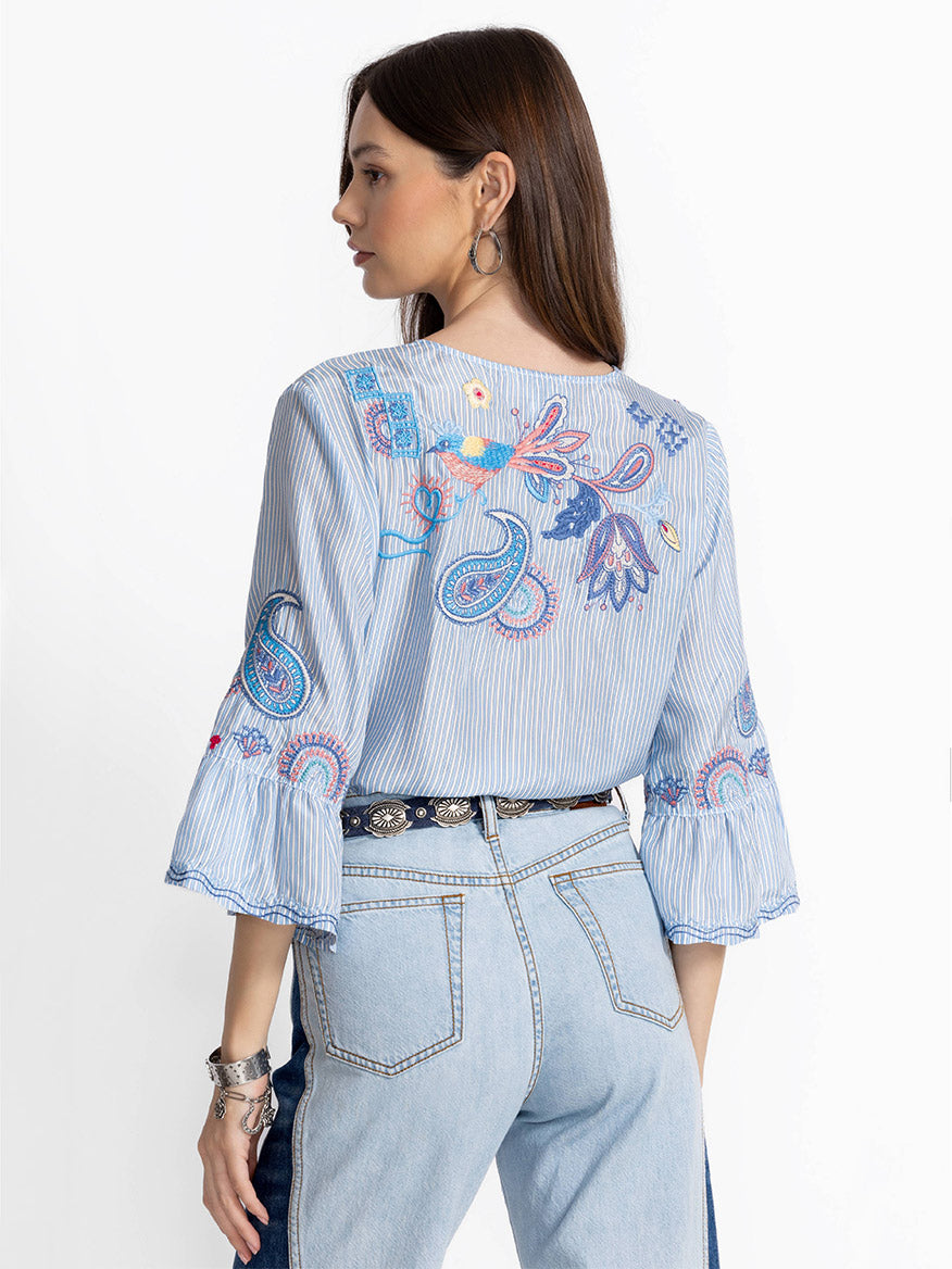 A woman wearing a Johnny Was Ruffle Sleeve Blouse in Barine Stripe Bar with luxurious silk and embroidered details stands slightly turned. Her hair is down, accessorized with bracelets and hoop earrings, adding elegance to her casual jeans outfit.