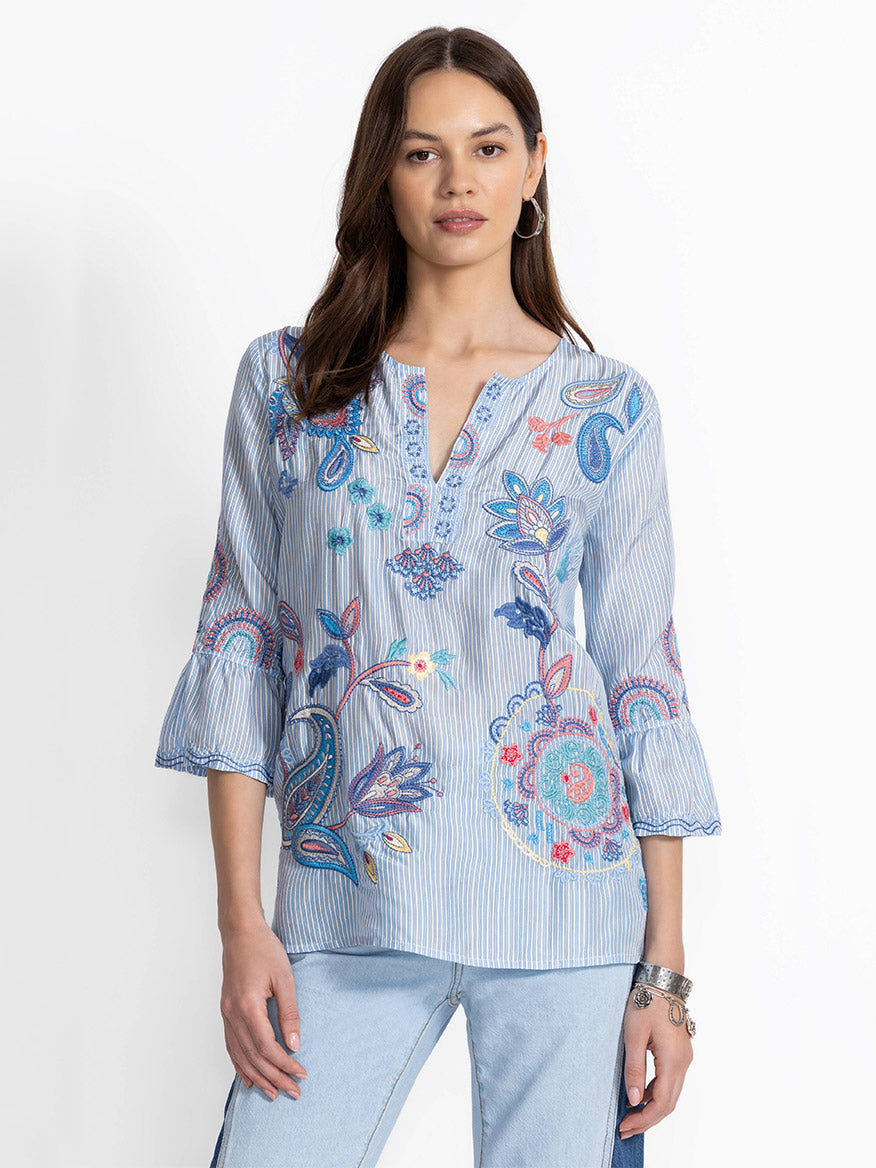 A woman wearing a Johnny Was Ruffle Sleeve Blouse in Barine Stripe, featuring floral embroidery and elegant ruffle sleeves, paired with light blue jeans, against a white background.