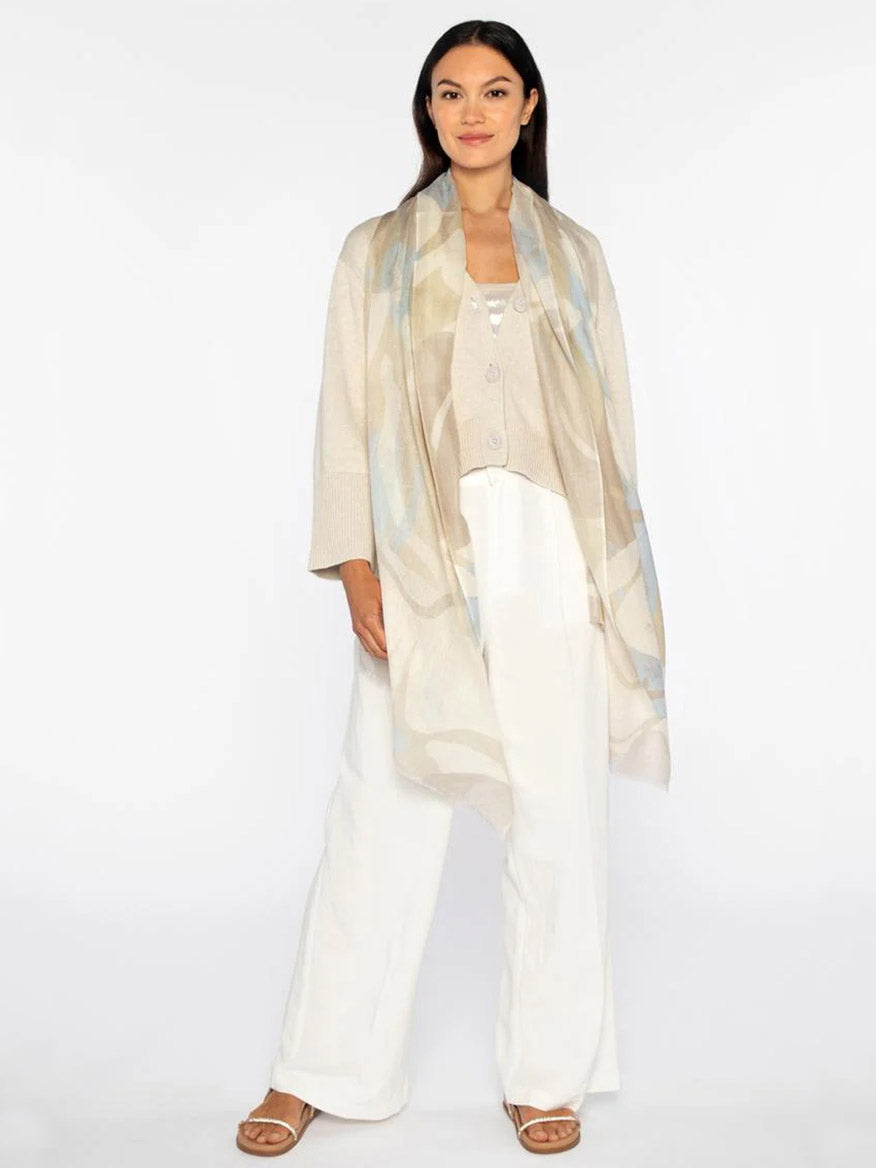 A woman stands elegantly against a plain background in a light beige long vest and white pants, her ensemble elevated by the Kinross Abstract Shadows Print Scarf in Almond Multi, adding sophistication to her pastel scarf and sandals.