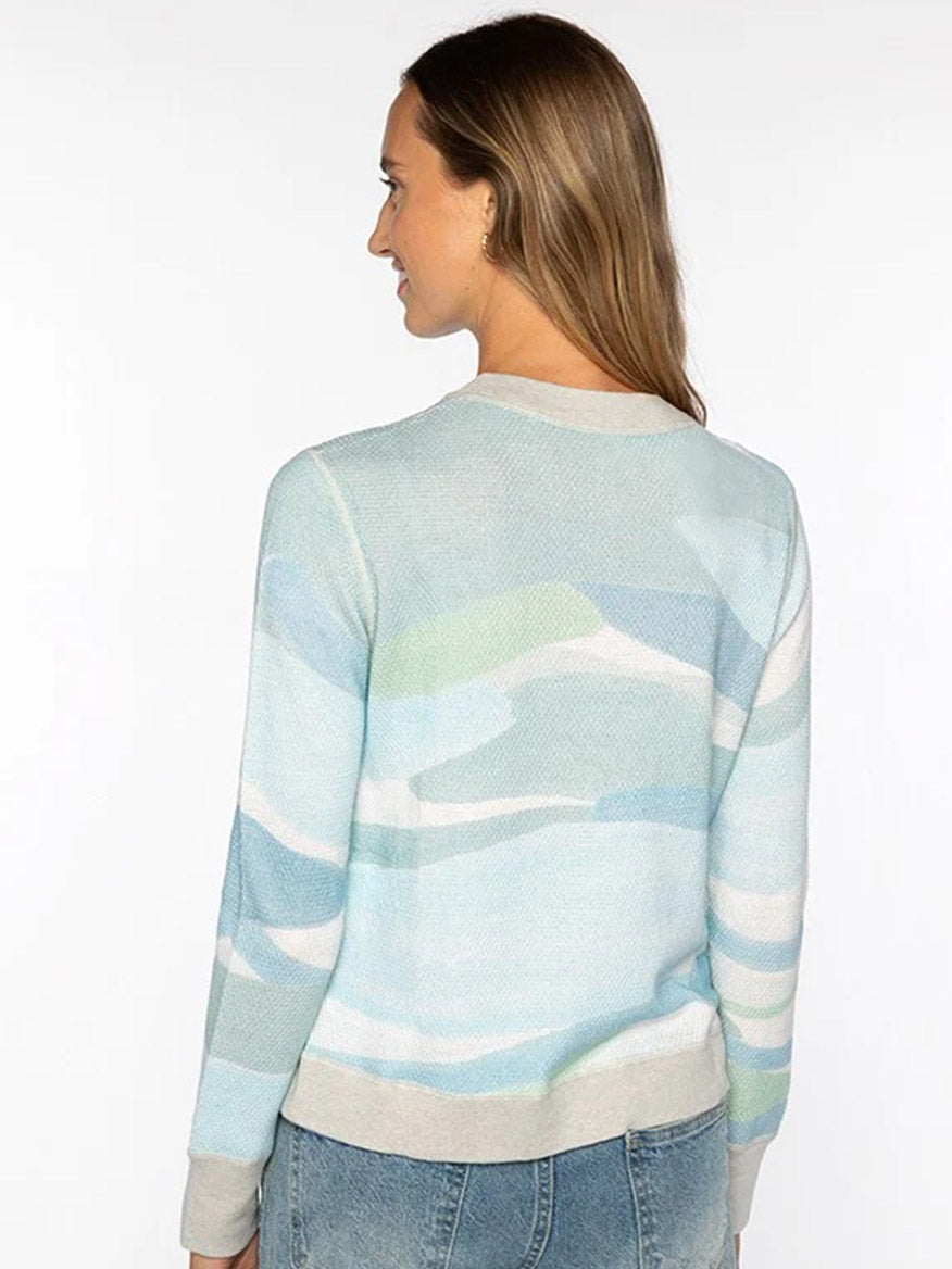 A woman with long brown hair, seen from the back, wears the Kinross Landscape Print Pullover in Multi—a soft cotton-cashmere blend in light blue and white—paired with blue jeans against a plain background.