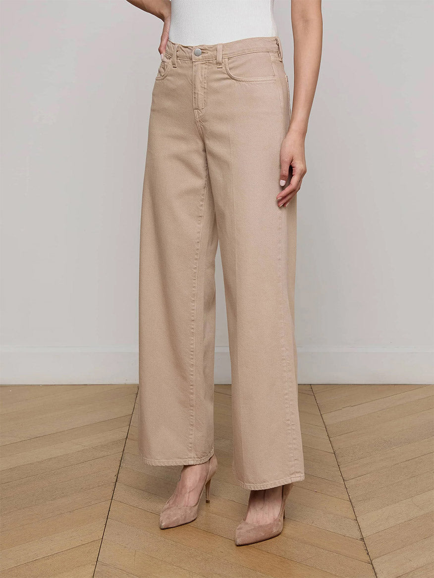 A person wearing L'Agence Alicent Wide-Leg Jean in Pale Khaki and beige heels stands on a wooden floor against a plain, light-colored wall.