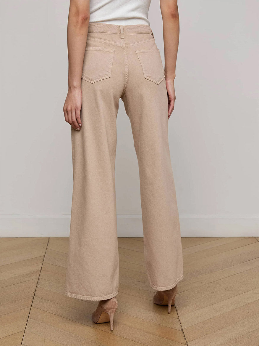 Wearing the L'Agence Alicent Wide-Leg Jean in Pale Khaki with nude heels, a person stands on a wooden floor, facing away from the camera against a plain wall.