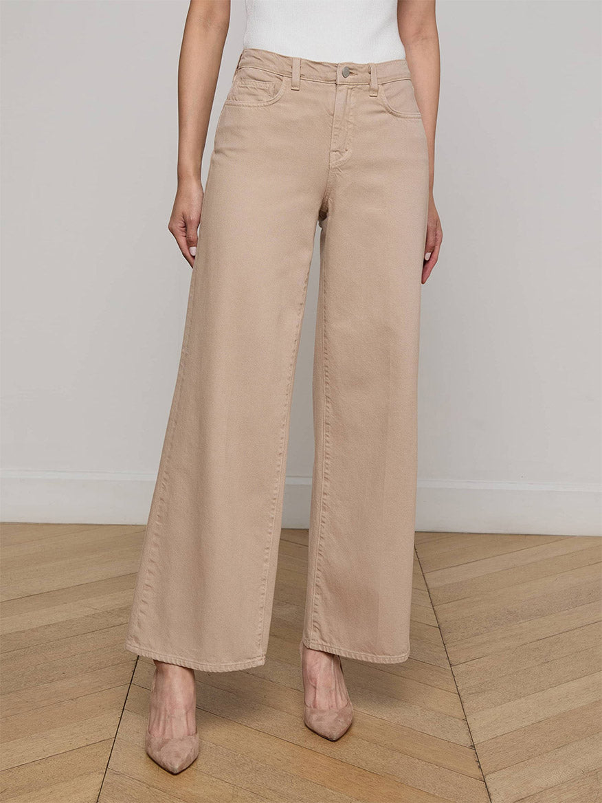 A person wearing the L'Agence Alicent Wide-Leg Jean in Pale Khaki and beige heels stands on a wooden floor against a plain light-colored wall.