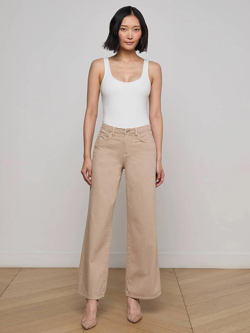 Woman standing indoors against a plain wall, wearing a white sleeveless top, the L'Agence Alicent Wide-Leg Jean in Pale Khaki, and beige heels.
