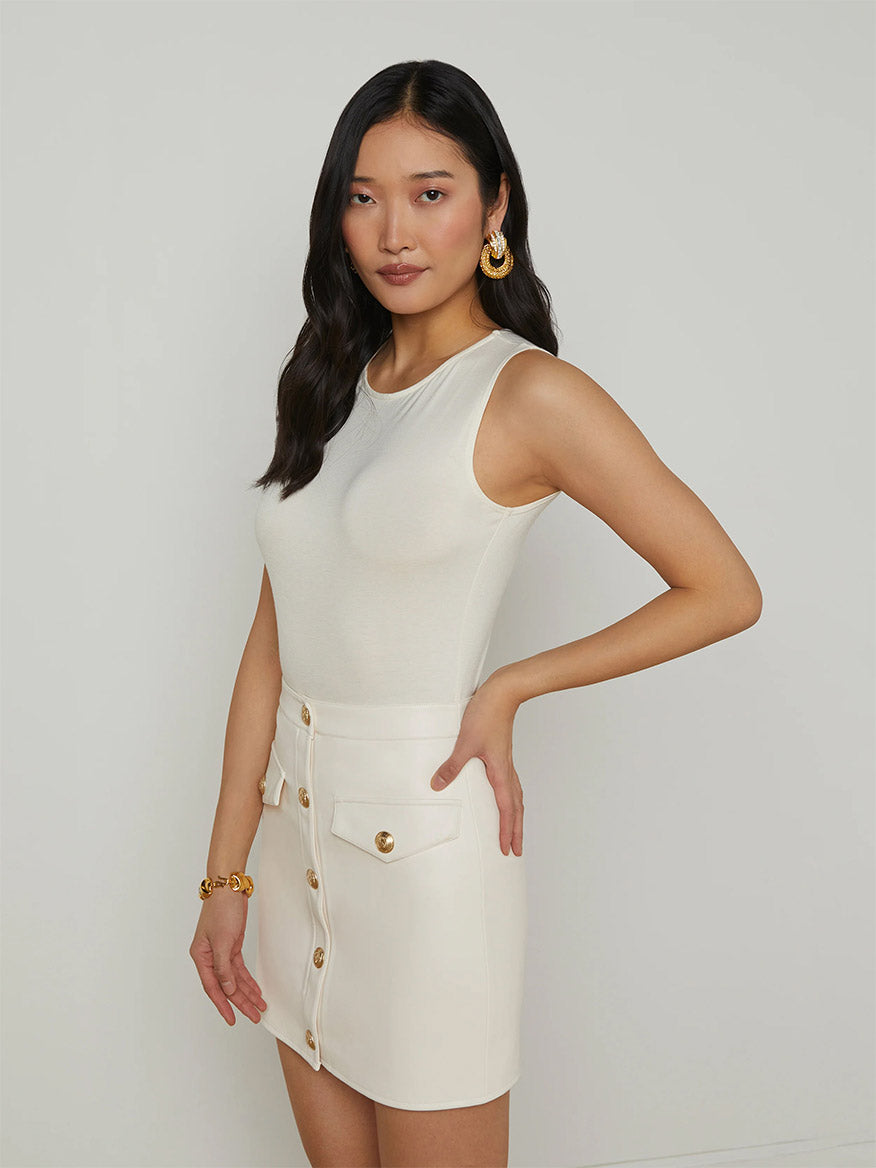 A woman stands against a plain background wearing the L'Agence Amia Tank Bodysuit in Vintage White and a high-waisted white skirt with front buttons.