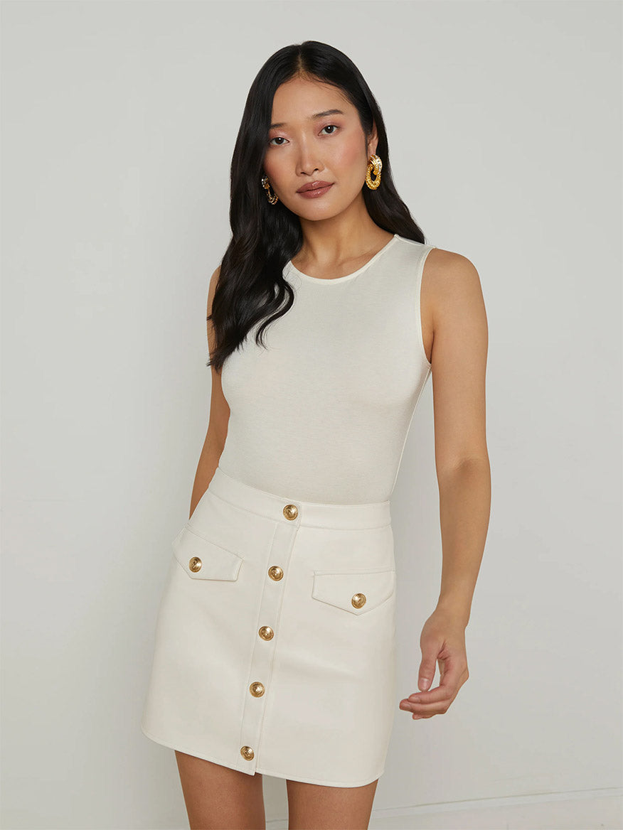 A woman wears the L'Agence Amia Tank Bodysuit in Vintage White paired with a white button-front skirt with gold buttons, standing against a plain light background.