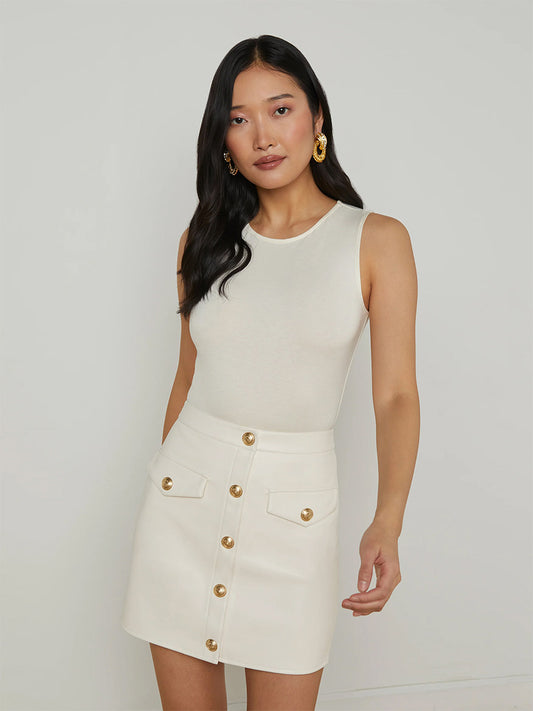 A woman wears the L'Agence Amia Tank Bodysuit in Vintage White paired with a white button-front skirt with gold buttons, standing against a plain light background.