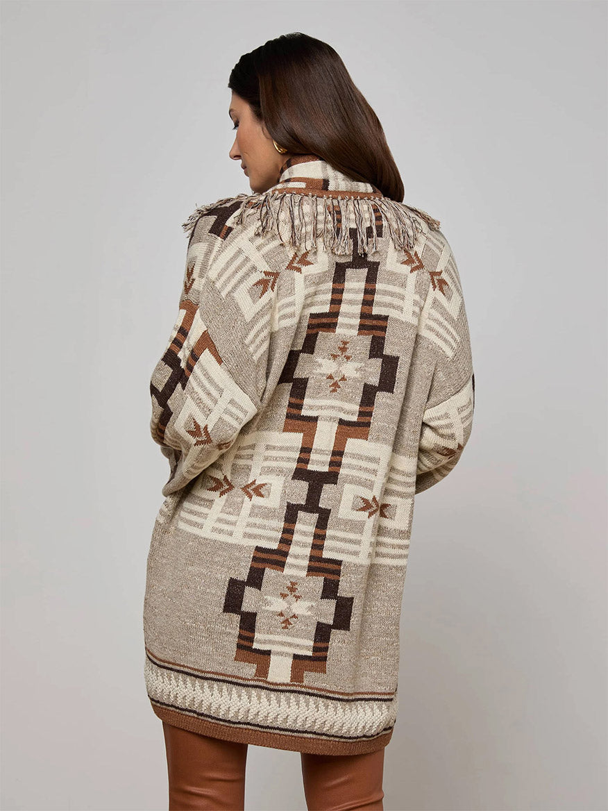 A woman stands facing away from the camera against a plain background, wearing the L'Agence Jair Cardigan in Natural Multi—featuring beige and brown cotton jacquard with geometric pattern and fringe detail on the back.