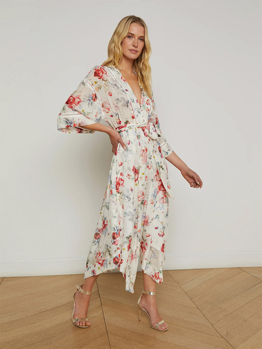 A woman stands indoors on a wooden floor wearing the L'Agence Miki Silk Dress in Ivory Multi Watercolor Dusty Floral, featuring a flowy silhouette, handkerchief hem, long sleeves, tie belt, and paired with strappy heels.