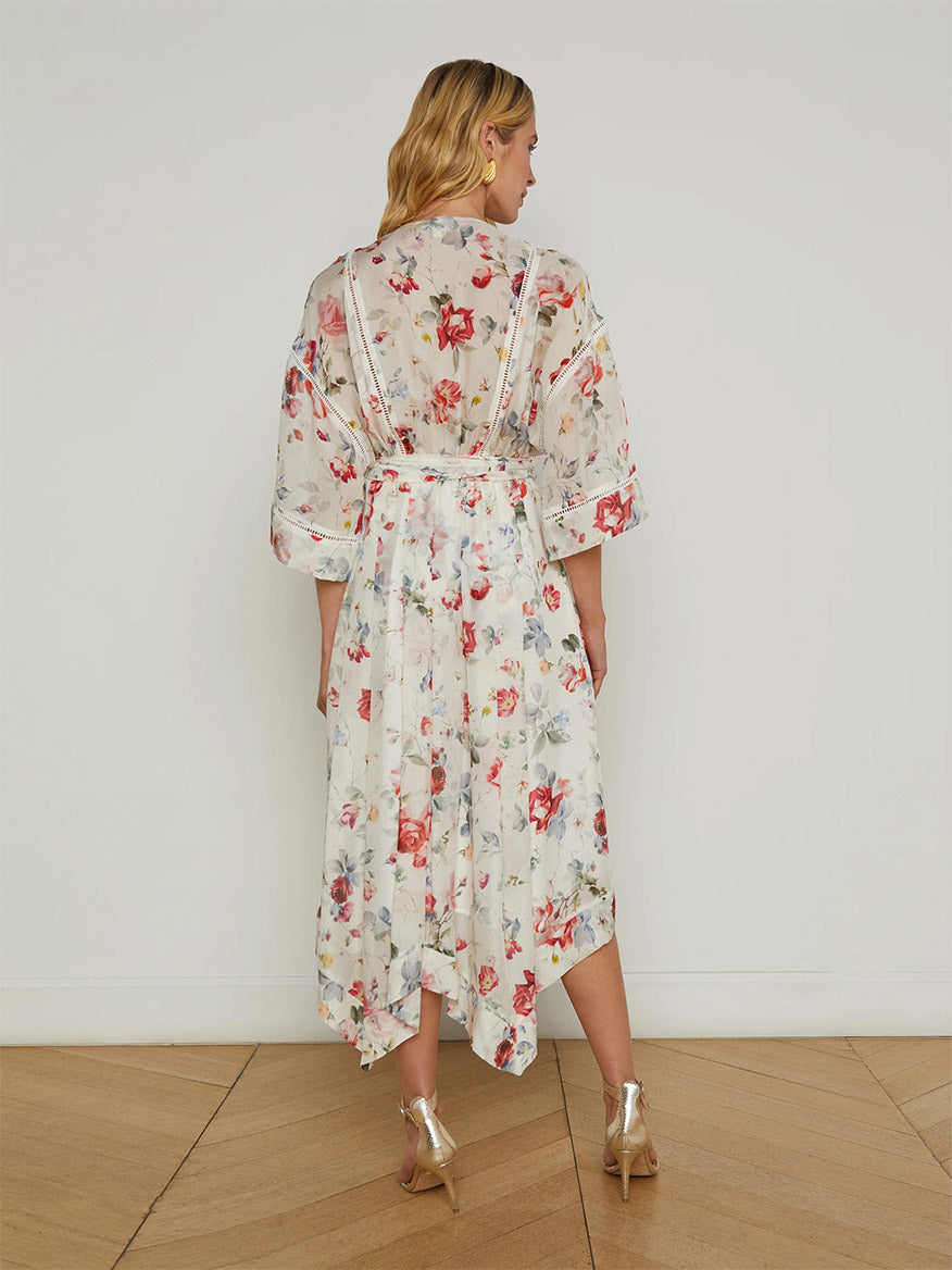 A woman faces away in the L'Agence Miki Silk Dress in Ivory Multi Watercolor Dusty Floral, featuring sheer sleeves and paired with gold heels, set against a plain white wall on a wooden floor.