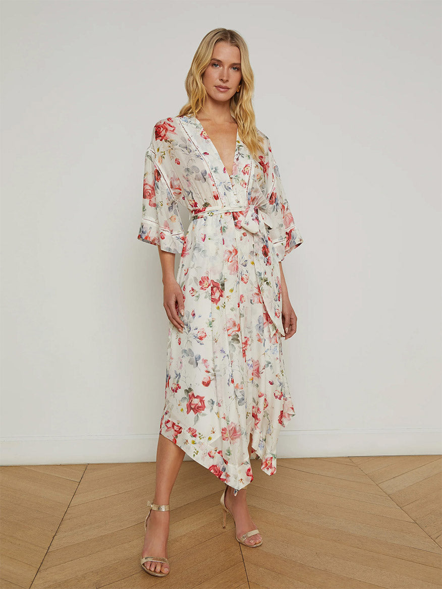 A woman stands indoors on a wooden floor wearing the L'Agence Miki Silk Dress in Ivory Multi Watercolor Dusty Floral, styled with gold heeled sandals and featuring a tie belt and graceful handkerchief hem.