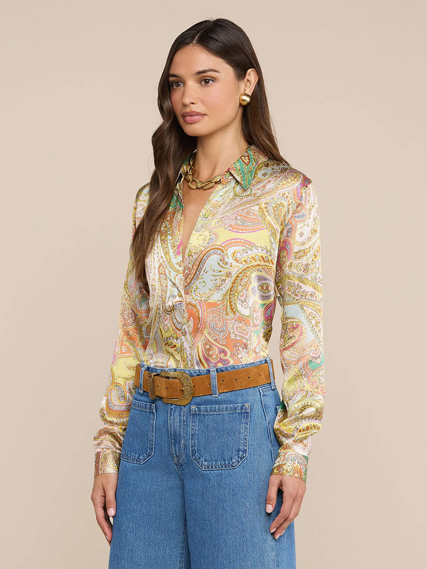 Woman wearing the L'Agence Tyler Blouse in Green Multi Cluster Paisley, paired with blue high-waisted jeans, a brown belt, gold earrings, and a gold necklace, standing against a plain beige background.