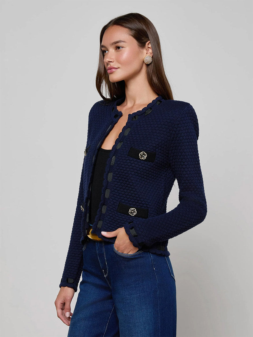 A woman wears the L'Agence Astor Cardigan in Midnight/Black, a textured knit with decorative buttons, over a black top and blue jeans, standing against a plain light background.