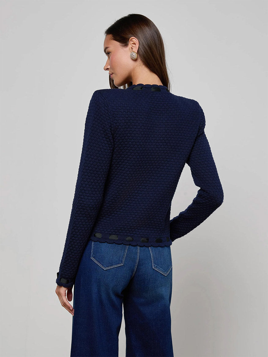 A woman faces away, wearing the L'Agence Astor Cardigan in Midnight/Black with blue jeans against a plain light background.