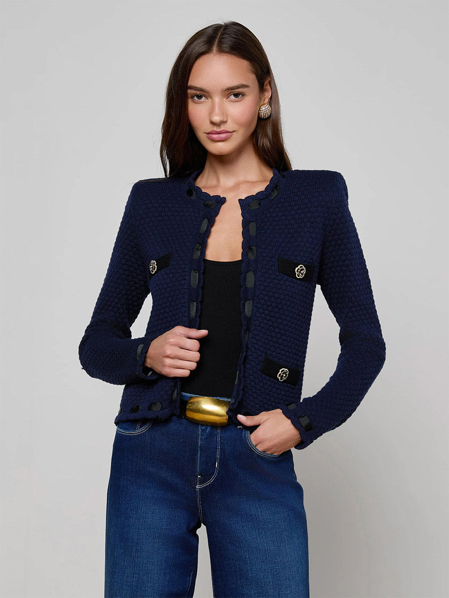 A woman wears the L'Agence Astor Cardigan in Midnight/Black, paired with a black top, blue jeans, and a wide gold belt, standing against a plain grey background.