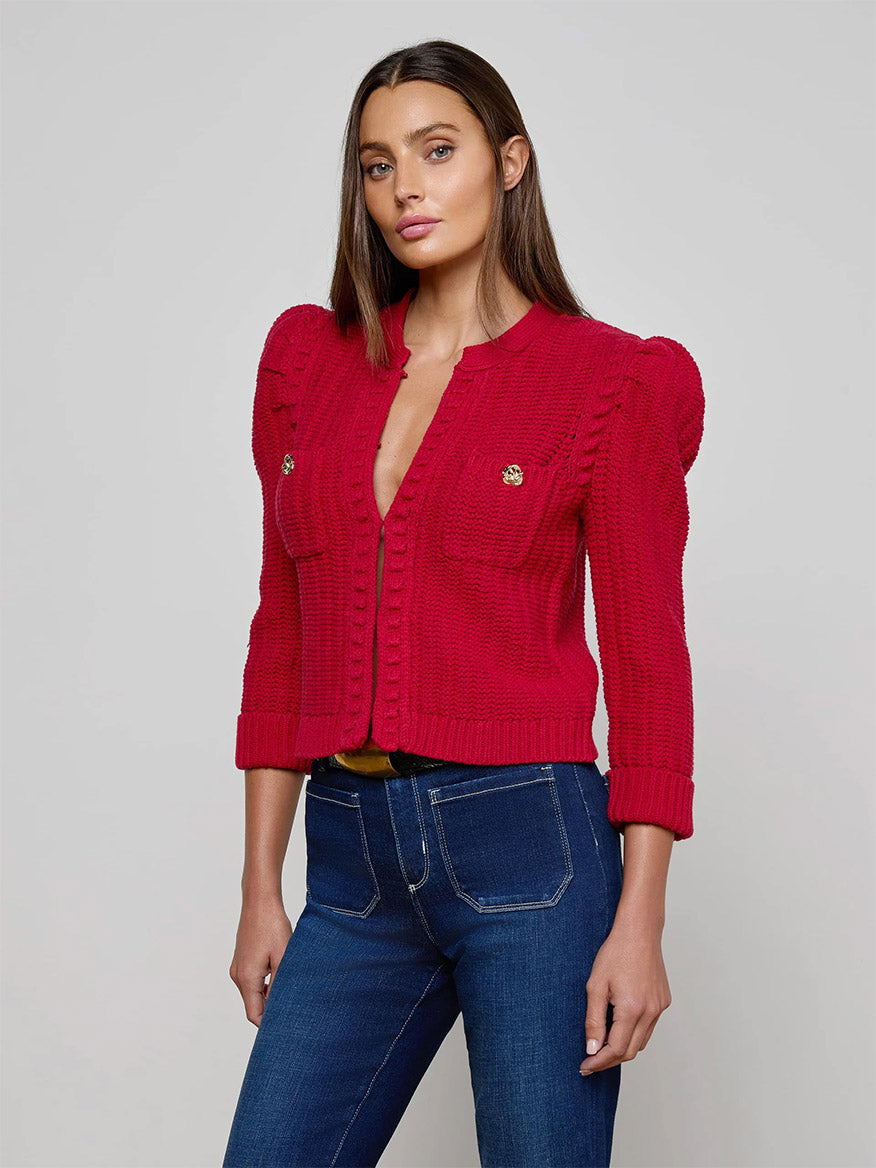 Woman wearing the L'Agence Delli Cropped Cardigan in Engine Red/Gold with puffed shoulders and blue jeans, standing against a plain light gray background.