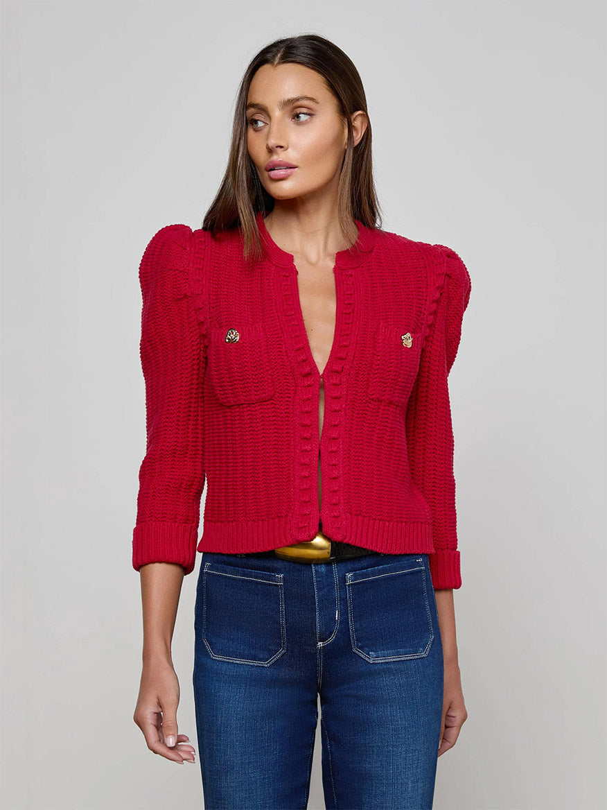 A woman wears the L'Agence Delli Cropped Cardigan in Engine Red/Gold, featuring textured knit, shoulder details, and front pockets, styled with blue jeans against a plain light gray background.