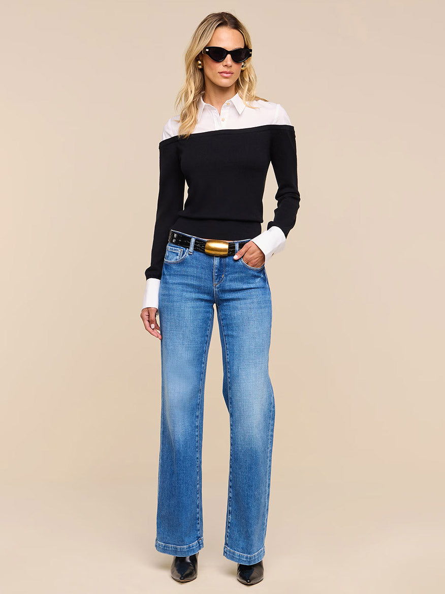 Woman wearing sunglasses, the L’Agence Mathilde Poplin Collar Pullover in Black/White, blue wide-leg jeans, a black belt, and black shoes standing against a plain beige background.