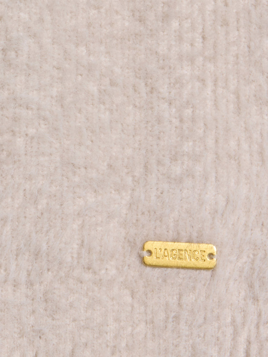 A small gold metal tag engraved with “L’AGENCE” is attached to a beige textured fabric background, evoking the relaxed sophistication and cloud-soft feel of the L’Agence Nimbus Pullover in Vintage White.