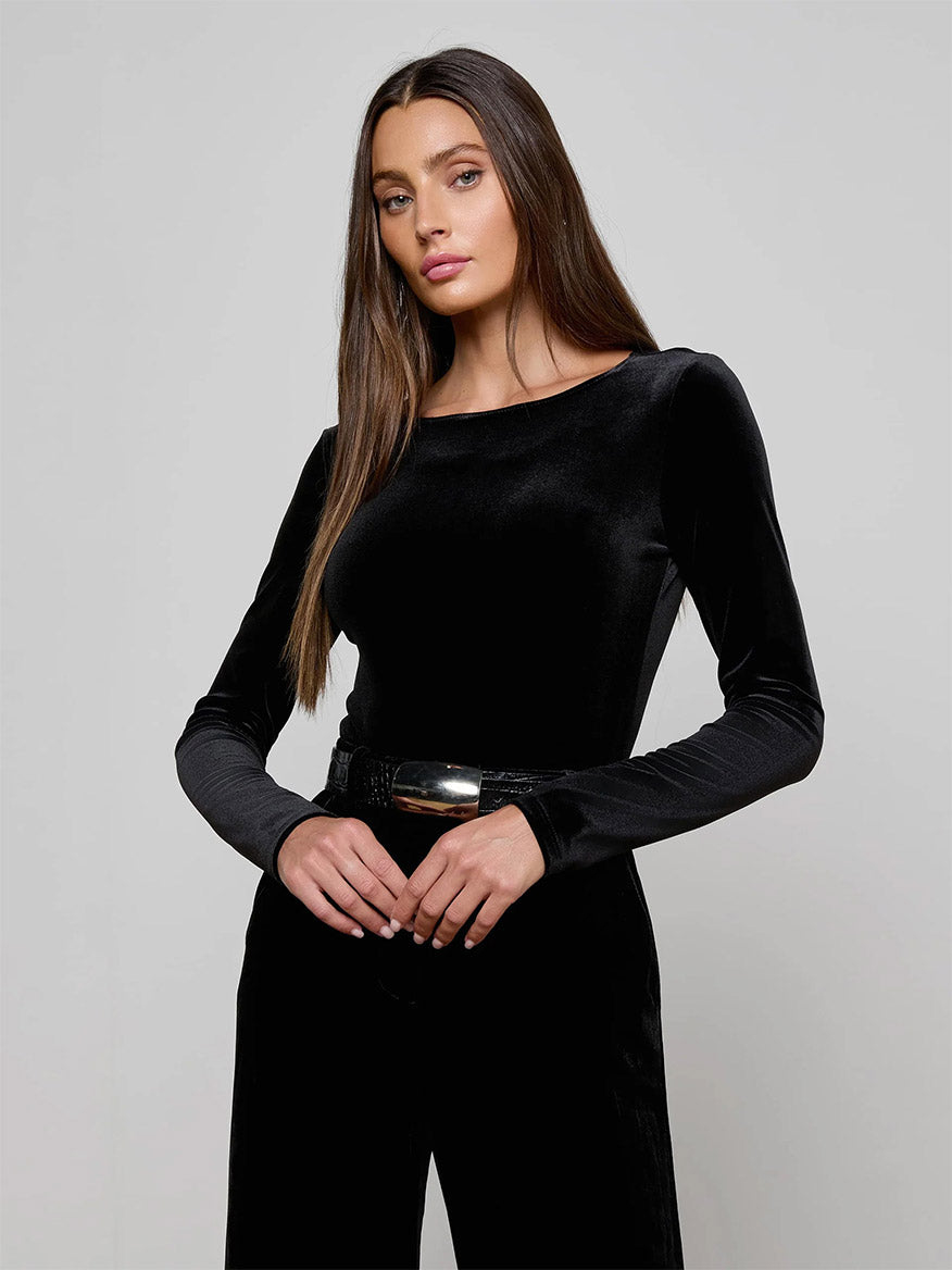 Woman with long brown hair wears the L'Agence Seraphine Velvet Bodysuit in Black and black pants, posing with hands clasped in front against a plain light background.