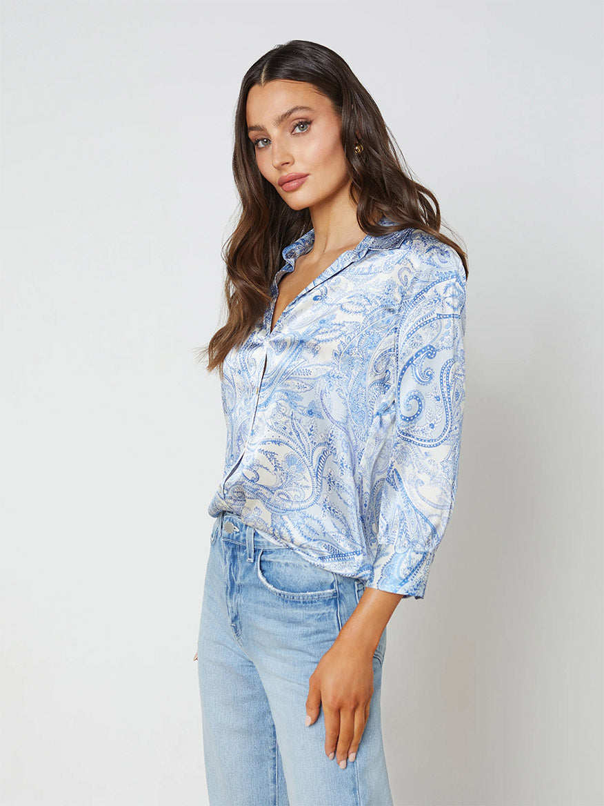 A woman in a L'Agence Dani Blouse in Ivory/Blue Decorated Paisley and light-wash jeans, posing against a plain background.