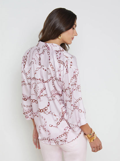 Woman viewed from the back wearing a L'Agence Patrice Blouse in Lilac Snow Multi Sketch Chain, paired with light pink trousers, standing against a white background.