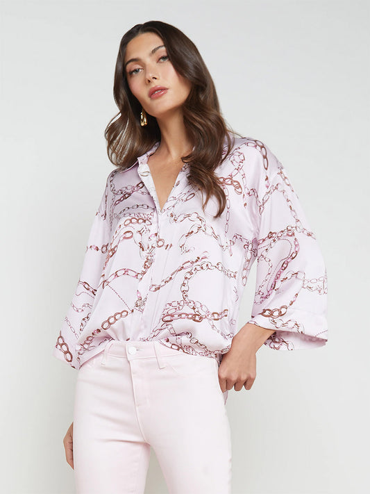 A woman models the L'Agence Patrice Blouse in Lilac Snow Multi Sketch Chain, a white pure silk top with pink chain motifs, paired with white pants in a relaxed-fit style, standing against a light grey background.