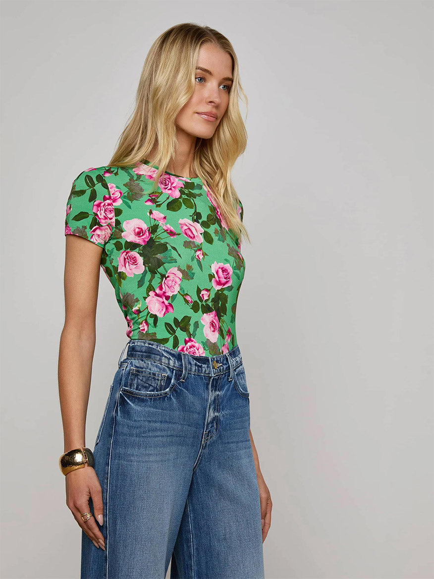 A woman with long blonde hair wears the L'Agence Ressi Fitted Tee in Green Multi Santa Fe Rose and high-waisted blue jeans, standing against a plain light background.