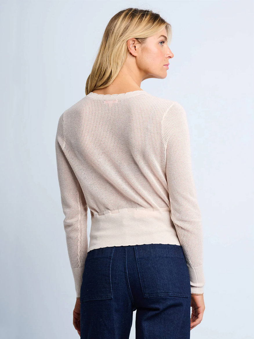 A woman with blonde hair faces away, wearing the Lisa Todd Flirt Cardigan in Sheepskin and dark blue jeans against a plain light background.